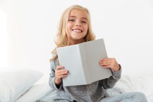 Cheerful little girl in gray pajamas holding book, looking at ca